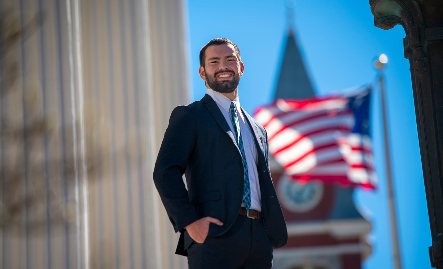 A UNCW student poses at Wilmington City Hall with an American flag and the spire of the New Hanover County Courthouse in the background